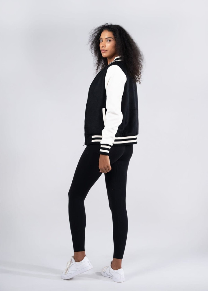 A black and white women's varsity jacket with a soft shell wool blend, modelled alongside black leggings and white shoes.