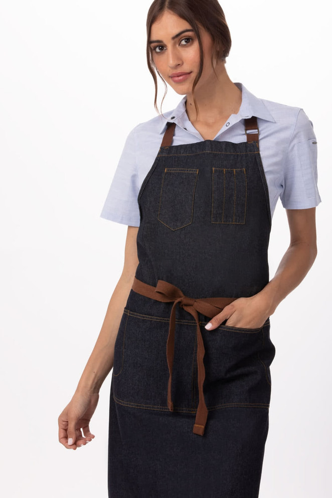 A denim apron in dark blue with a brown waist tie, featuring front pockets and a logo.