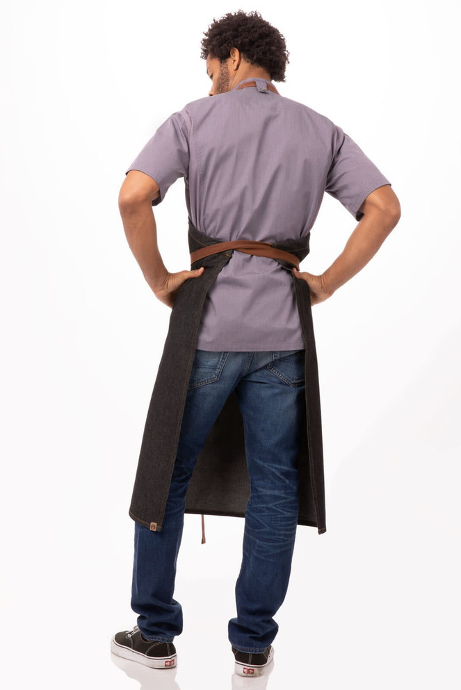 A dark grey bib apron worn by a person facing away, with a brown belt, styled over a grey shirt and blue jeans.