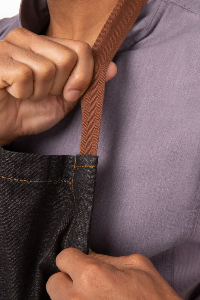 A close-up of a person holding the brown strap of a dark denim chef's bib apron. The background is neutral.