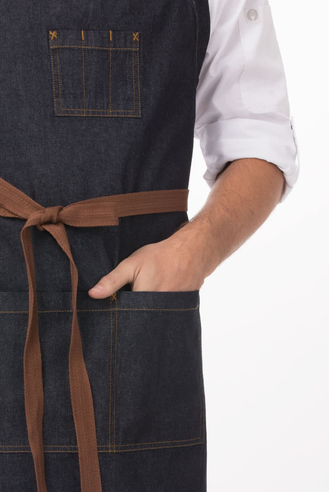 A dark denim apron with a brown tie, featuring a pocket and stitching details, models a stylish workwear look.