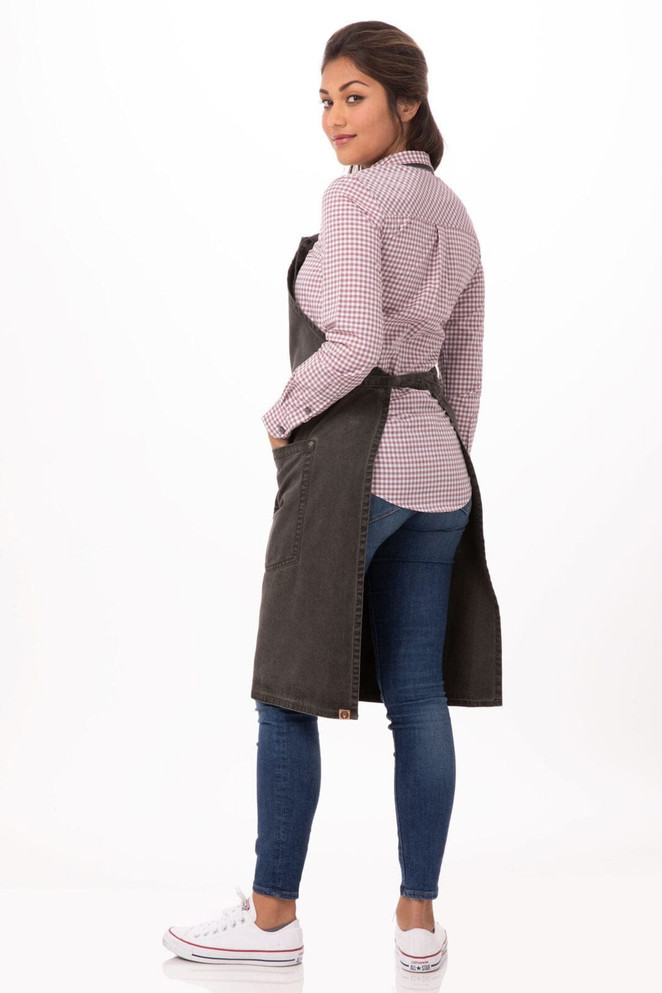 A Dorset Bib Apron in a dark colour, worn over a checked shirt, paired with blue jeans and white sneakers.