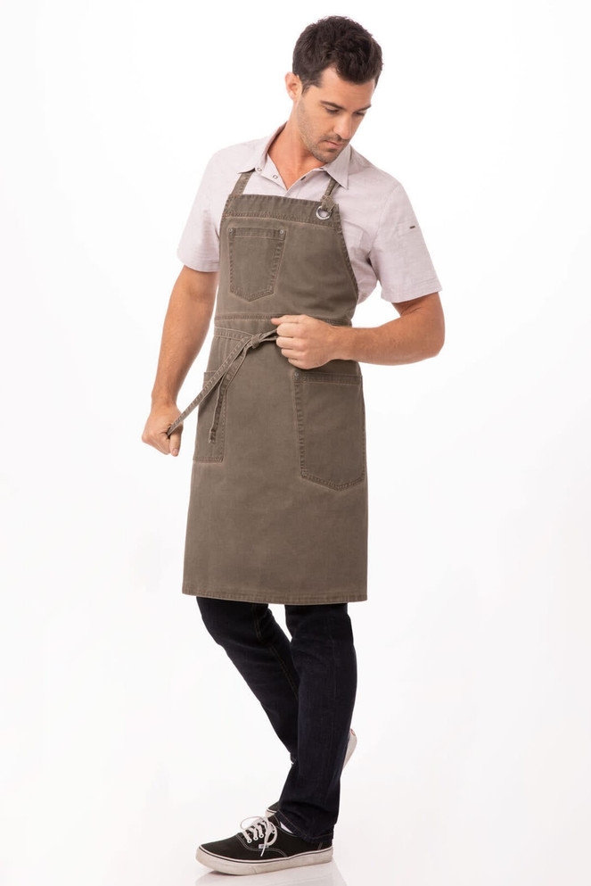 A man wearing a dark green Dorset Bib Apron with a front pocket, styled with a shirt and dark jeans.