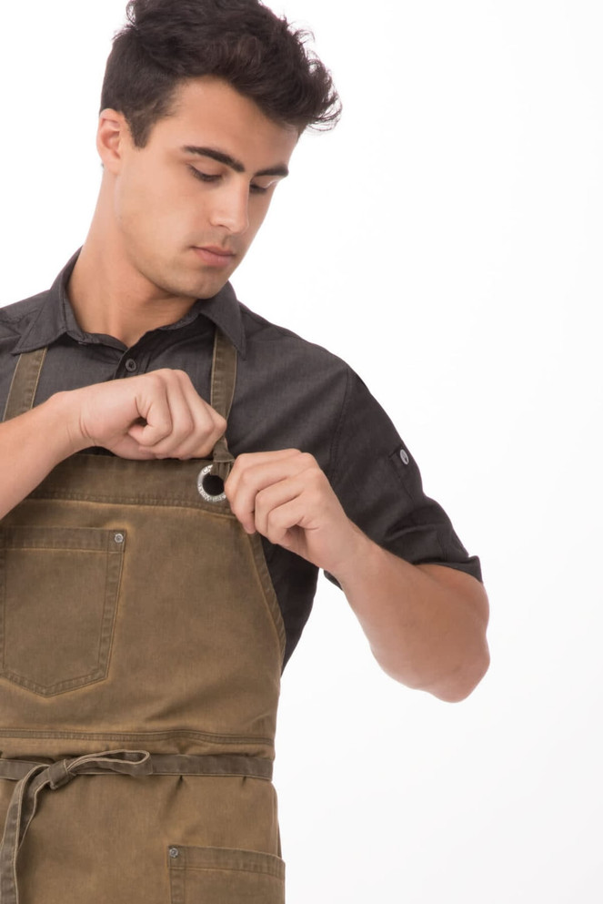 A man adjusts a green Dorset bib apron, featuring pockets and a logo, while wearing a black shirt.