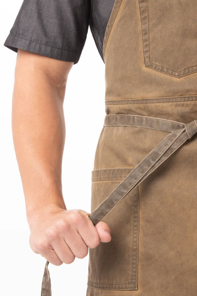 A person in a dark shirt holds the tie of a brown Dorset Bib Apron featuring two front pockets and a logo.