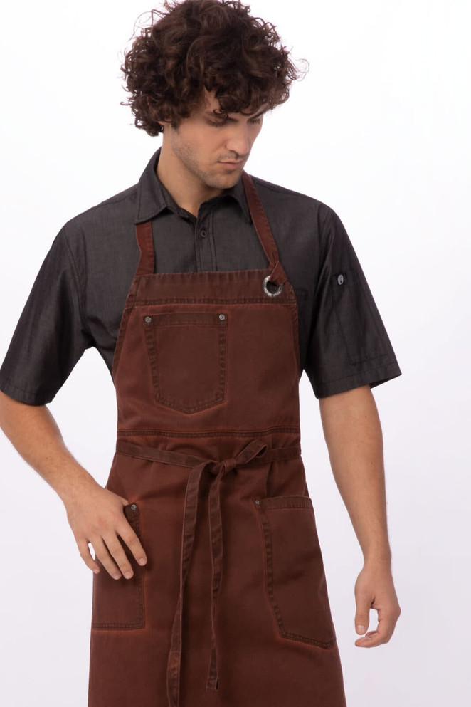 A brown bib apron with two pockets and adjustable straps worn by a model. The model is also wearing a black shirt.