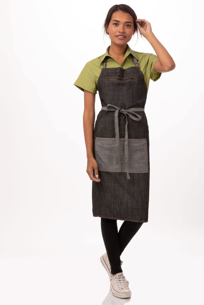 A woman in a green shirt and black Manhattan bib apron stands against a white background. The apron features a front pocket and ties.