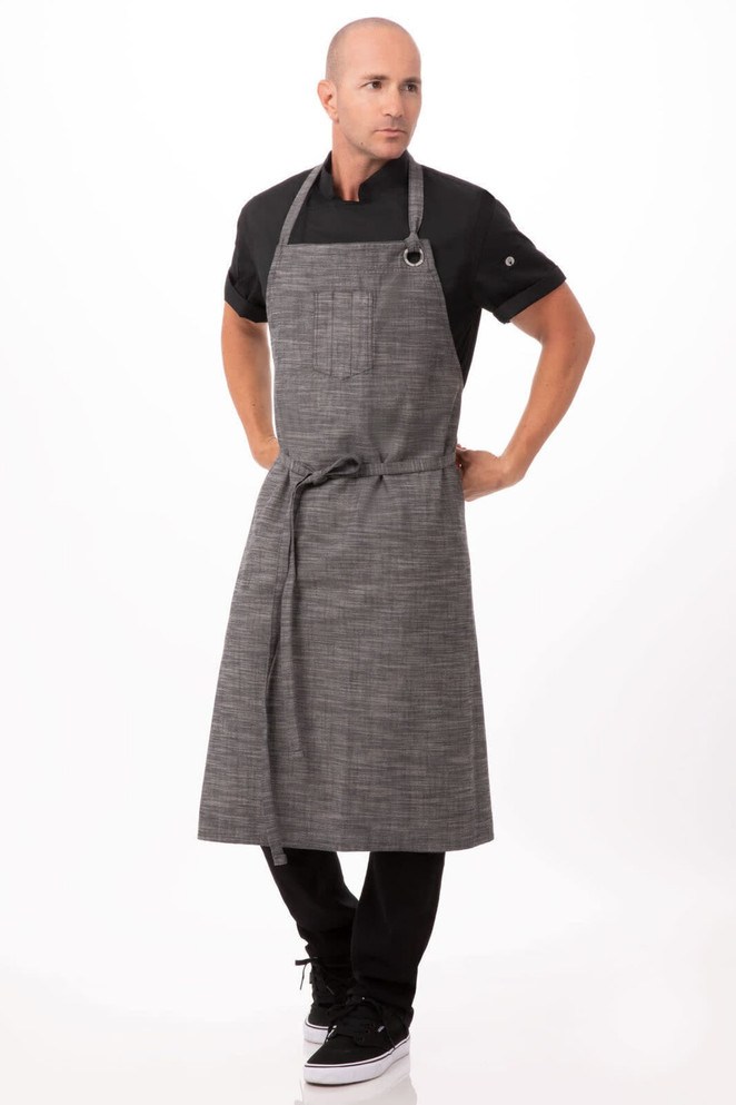 A man in a grey and black checkered bib apron poses against a white background, wearing a black shirt and dark pants.