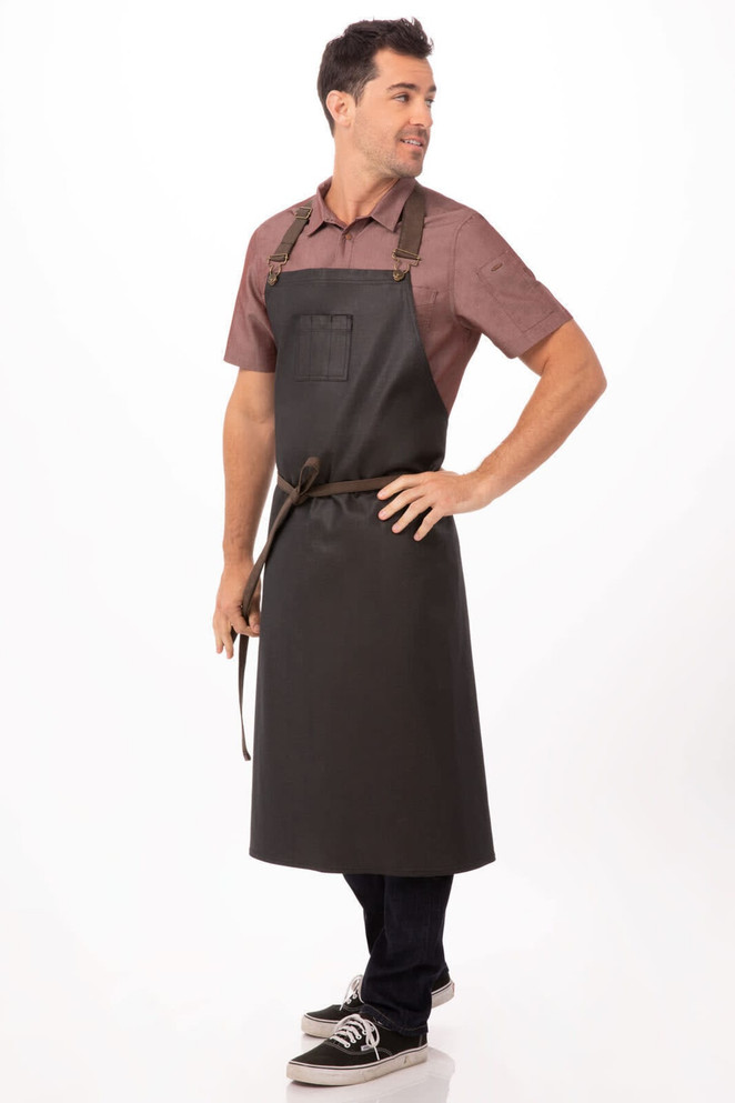 A dark grey chef's bib apron with adjustable straps, featuring a front pocket and a waist tie, worn by a model.