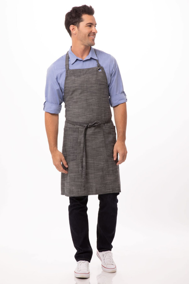 A man wearing a grey bib apron over a blue shirt, styled with black trousers and white shoes. The apron features a logo.