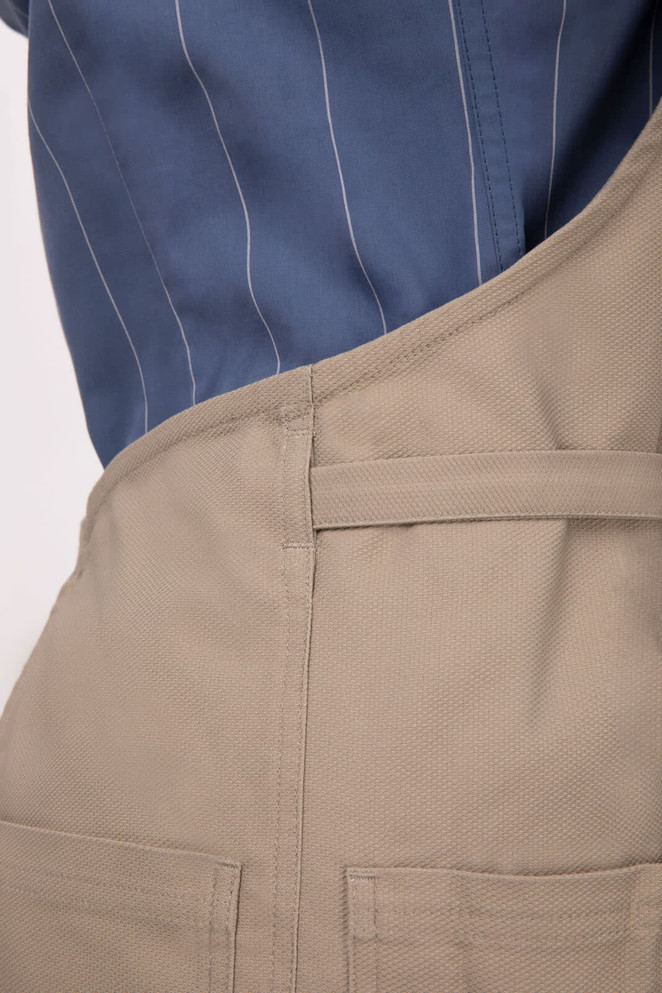 A beige Logan Bib Apron featuring pockets, worn over a blue pinstripe shirt, with a logo visible.