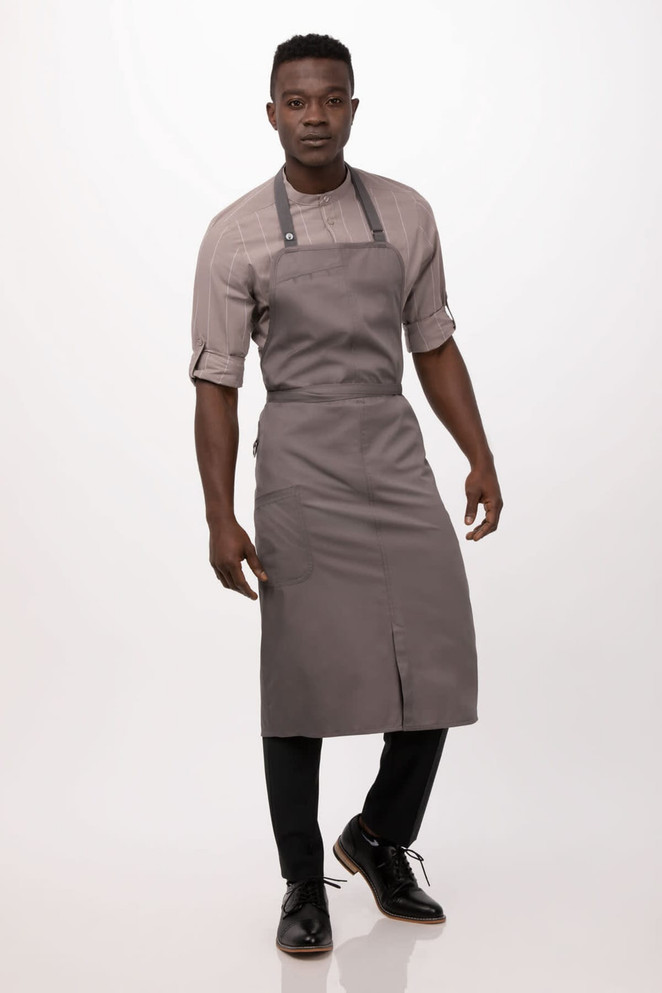 A grey bib apron worn over a light-coloured shirt, complemented by black trousers and shoes. The model stands confidently.