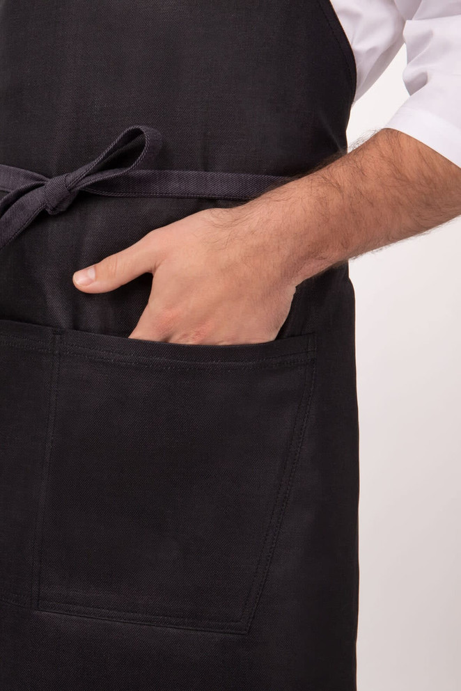 A charcoal apron featuring a waist tie and a front pocket, with a hand positioned inside the pocket.