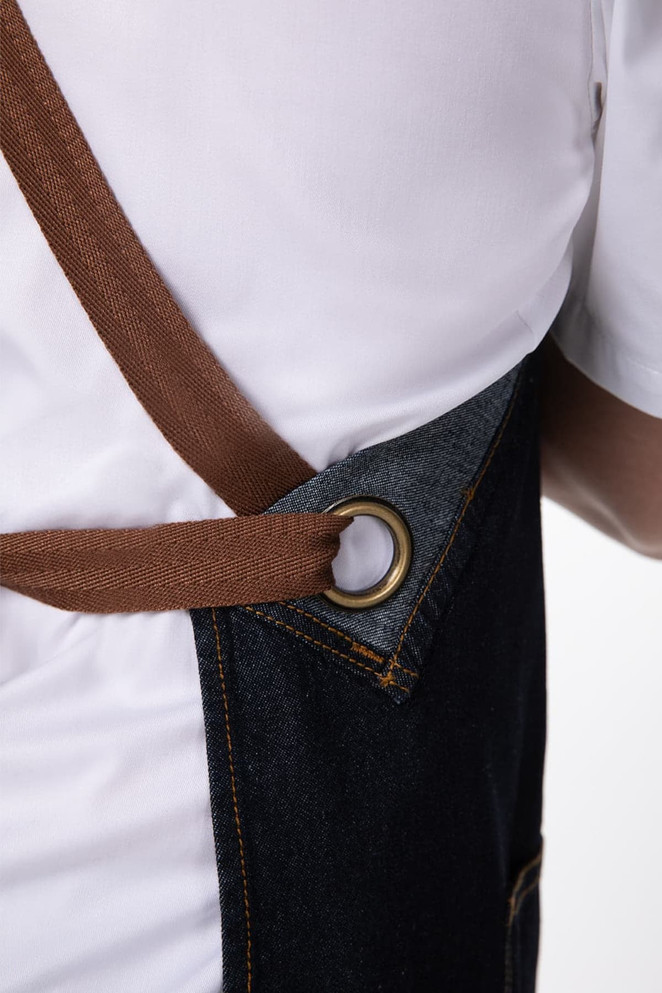 A denim cross back apron featuring a brown strap and a metal ring detail, demonstrating its design and material.