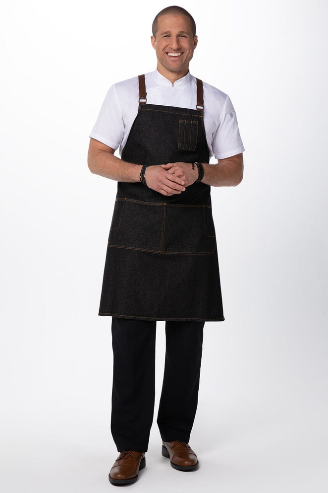 A man wearing a black denim cross back apron with brown straps, complemented by a white shirt and dark pants.