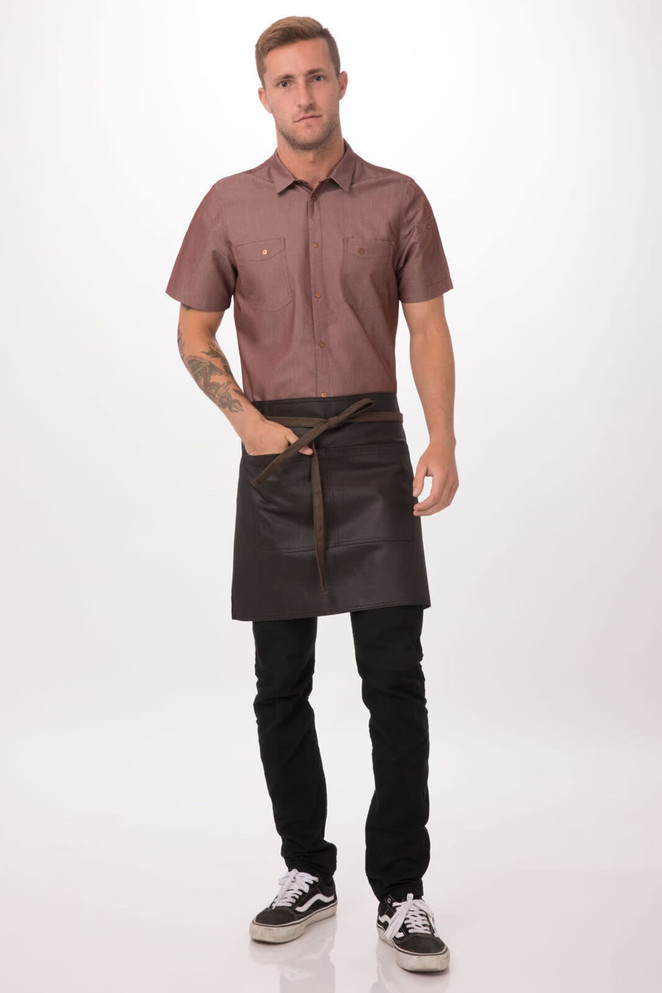 A man wearing a brown shirt and a black half apron with a waist tie, standing against a light background.