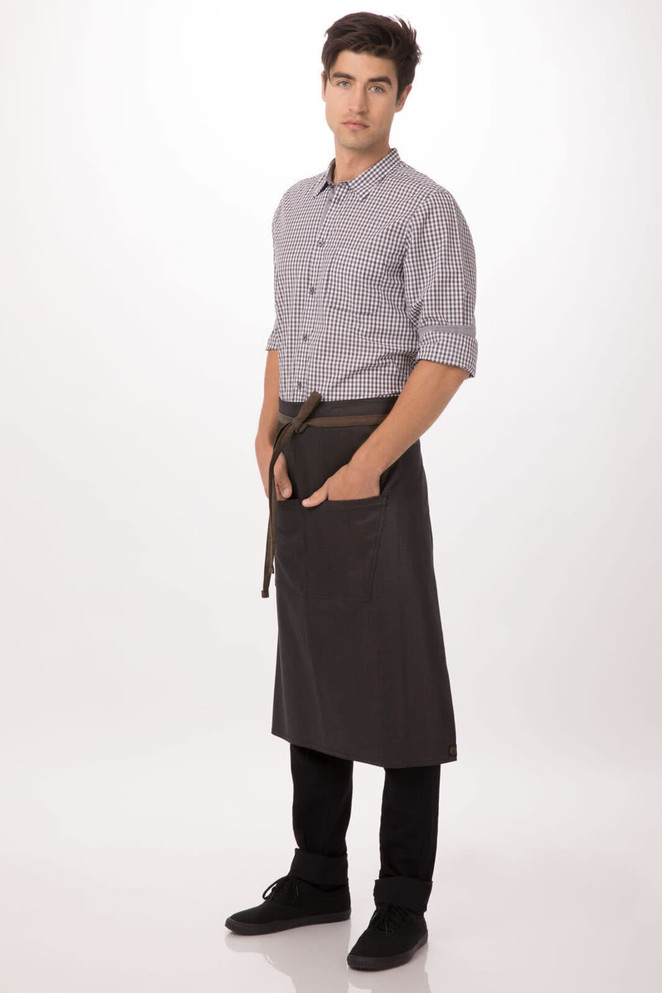 A man wears a dark charcoal Boulder Bistro Apron over a checked shirt, featuring pockets and a tied waist.