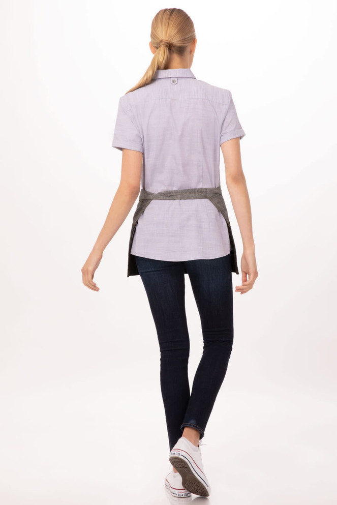 A waist apron in grey, worn over a light purple shirt, paired with dark jeans and white sneakers. It features a logo.