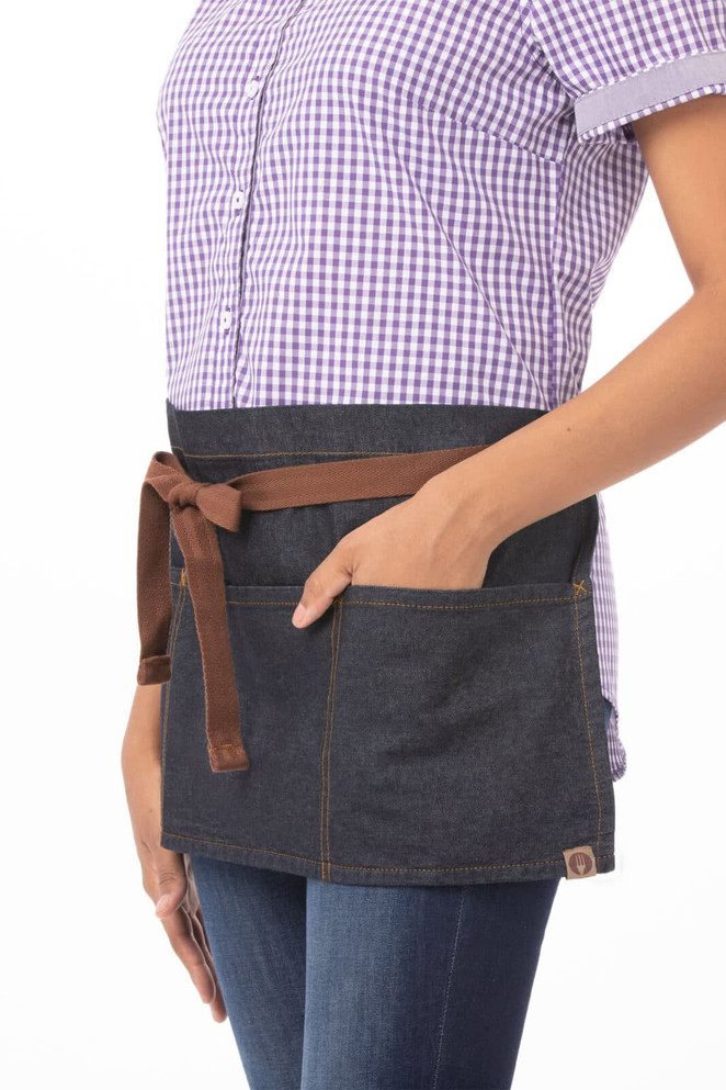 A waist apron in dark denim with brown leather straps and pockets, worn by a person in a checkered shirt.