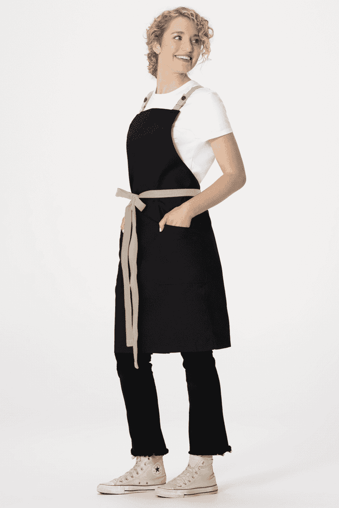 A black cross back apron with a beige belt and pockets, worn by a model with curly hair.