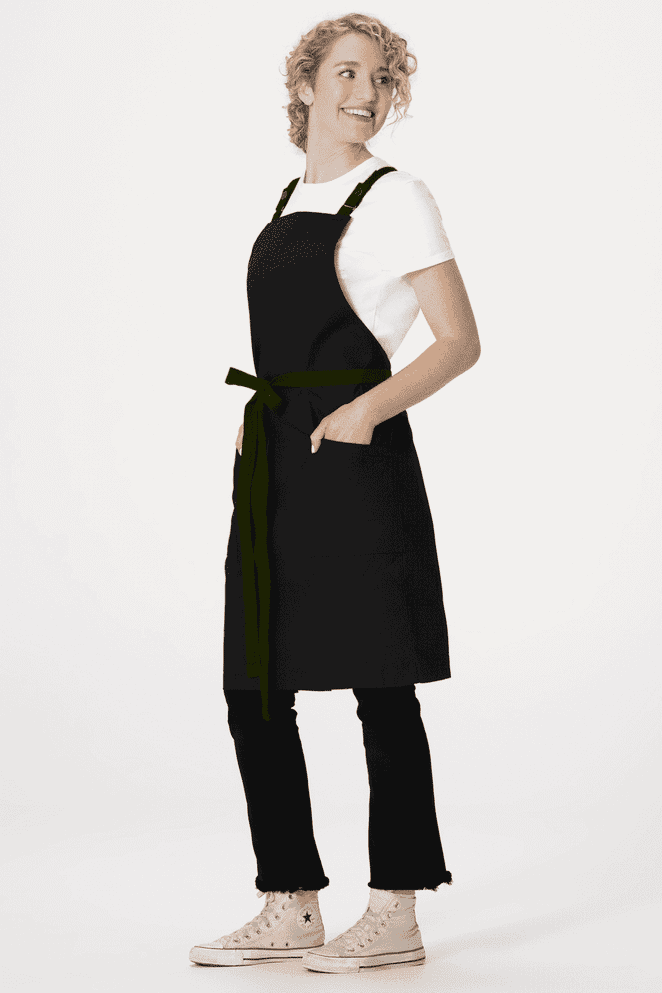 A black cross back apron with pockets worn over a white shirt, modelled by a person standing in profile.