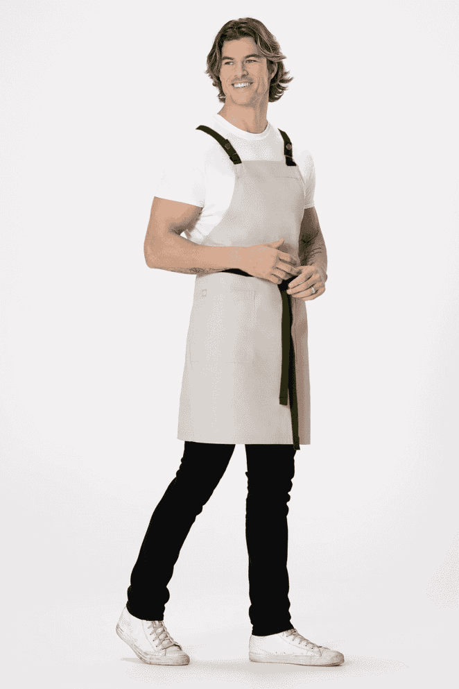 A light grey cross back apron worn by a model, featuring black straps, paired with black pants and white shoes.
