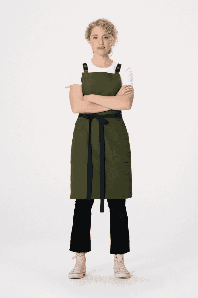 A green Lockharte cross back apron with black straps, worn by a model standing with arms crossed.