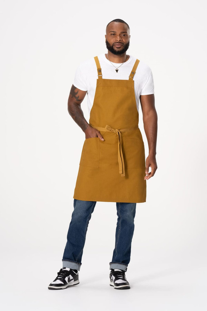 A man wearing a mustard yellow cross back apron, paired with a white t-shirt and blue jeans, standing hands on hips.