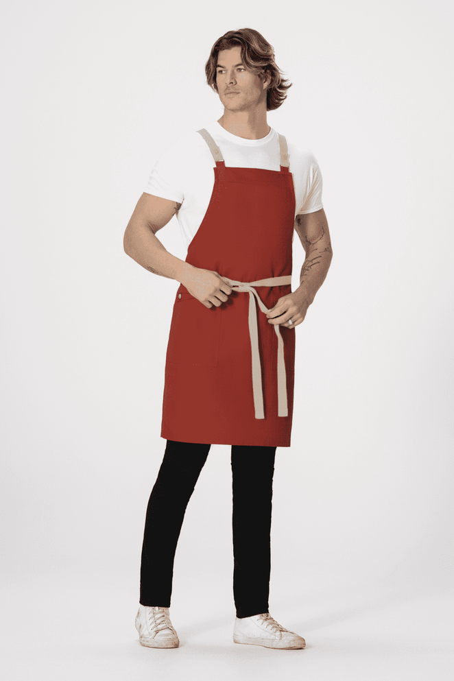 A model wearing a red Lockharte Cross Back Apron with grey straps, paired with a white t-shirt and black pants.
