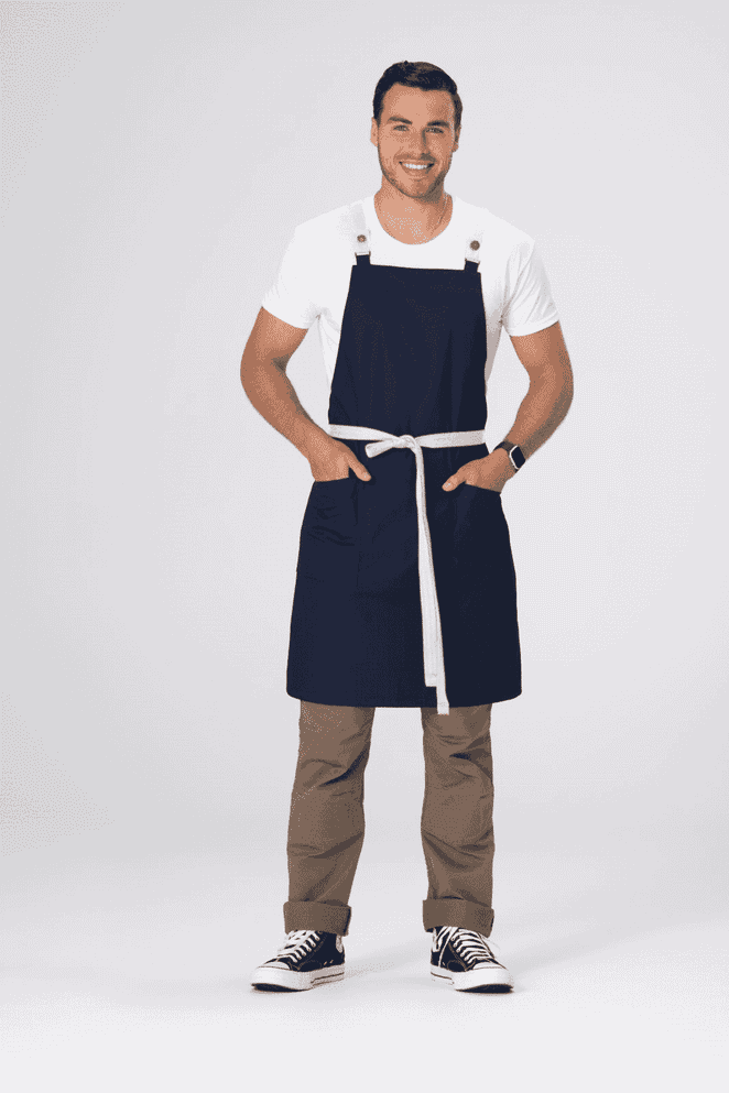 A man wearing a navy blue Lockharte Cross Back Apron with a white belt, standing confidently with hands in pockets.