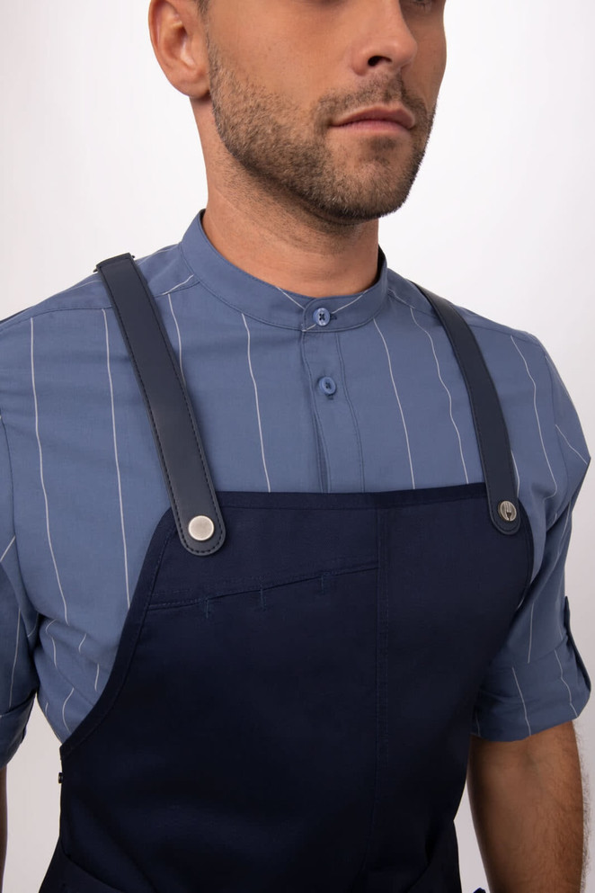 A navy blue bib apron with leather straps worn over a light blue striped shirt. Includes a logo detail.