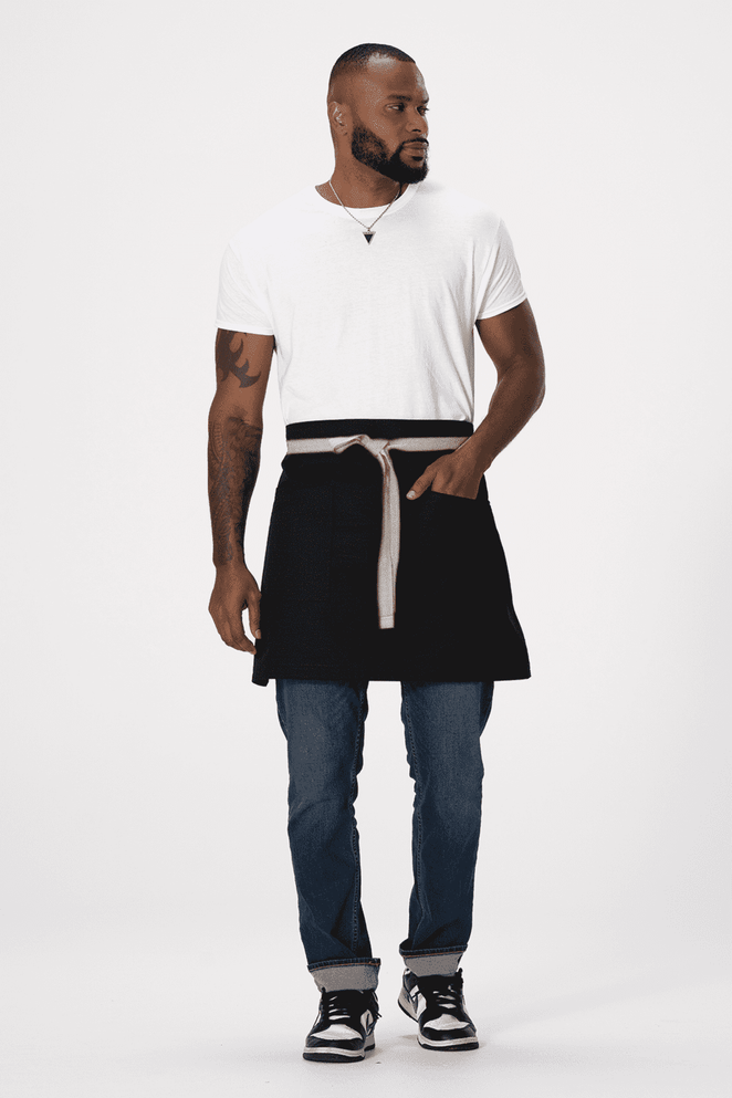 A man wearing a black Lockharte waist apron over a white T-shirt, paired with blue jeans and sneakers. The apron features a logo.