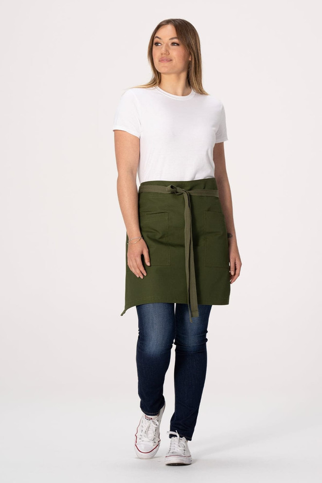 A woman wearing a green Lockharte waist apron over a white t-shirt and blue jeans, with pockets and a tie belt.