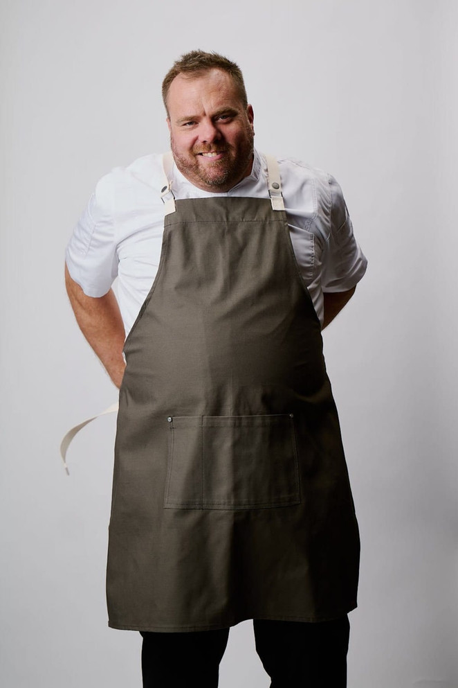 A man in a khaki cross-back apron smiles, showcasing its practical design and features. The apron includes a logo.