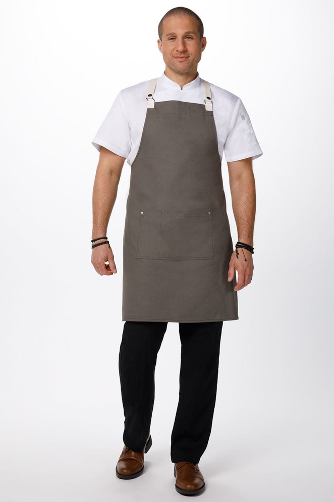 A man wearing a grey Byron Cross-Back Apron over a white shirt, standing with hands at his sides. The apron features a logo.