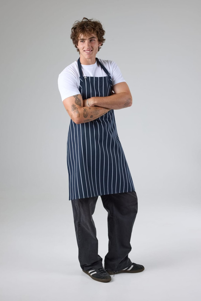 A navy and white striped bib apron worn by a smiling model, paired with a white t-shirt and dark trousers.