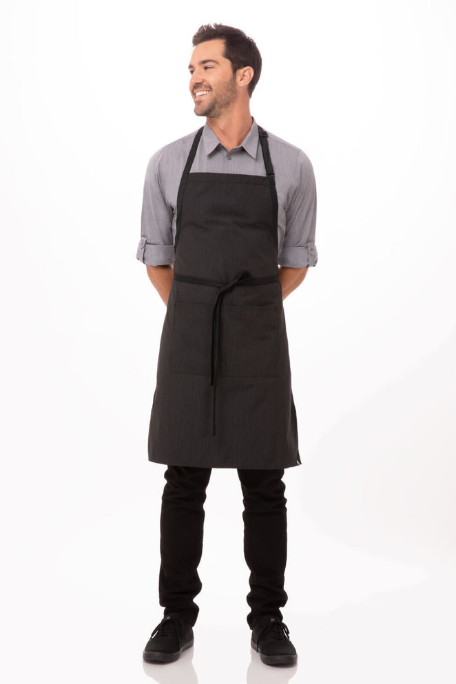 A black bib apron with contrasting ties worn by a smiling man, standing upright with hands behind his back.