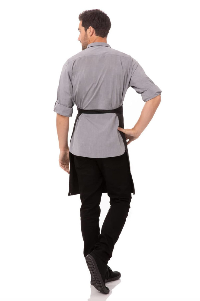 A man wearing a grey shirt and black bib apron with contrasting ties, standing with his back to the camera.
