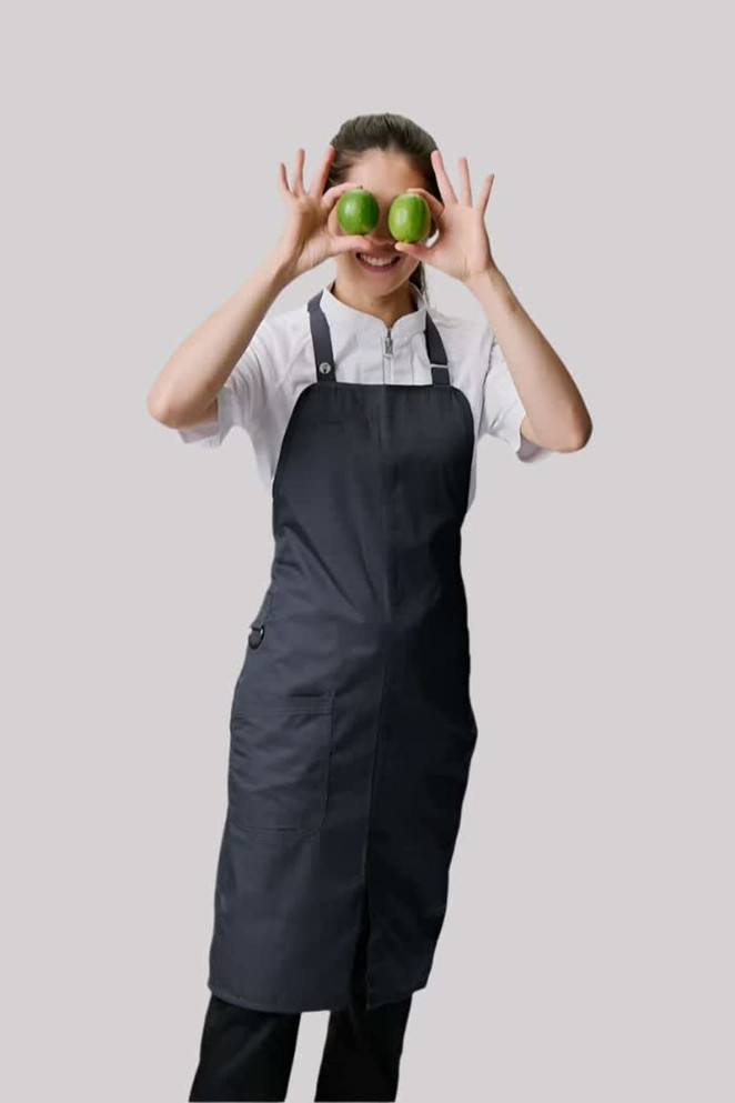 A dark grey bib apron worn by a person holding two green apples, smiling playfully. The apron features adjustable straps.