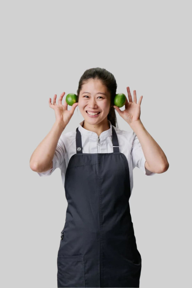 A woman in a black bib apron holds green limes to her ears, smiling against a neutral background.