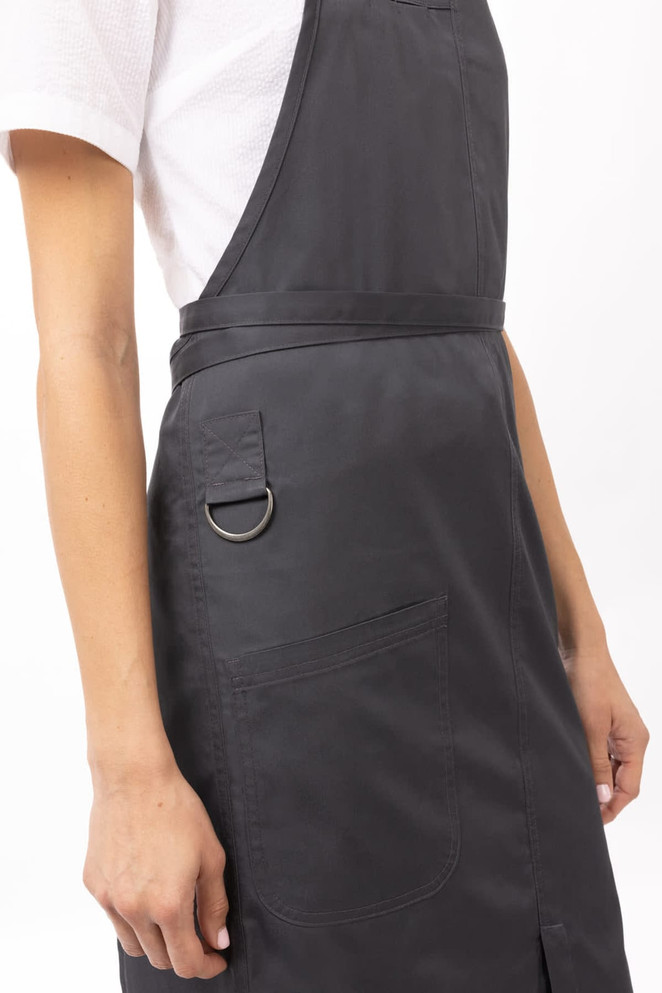 A black bib apron with a pocket and a metal ring detail, worn by a person against a neutral background.