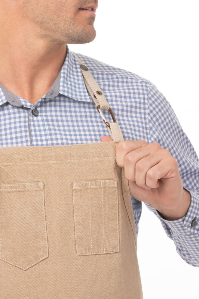 A beige bib apron with pockets, worn by a person in a blue checked shirt. The apron features a logo on the strap.