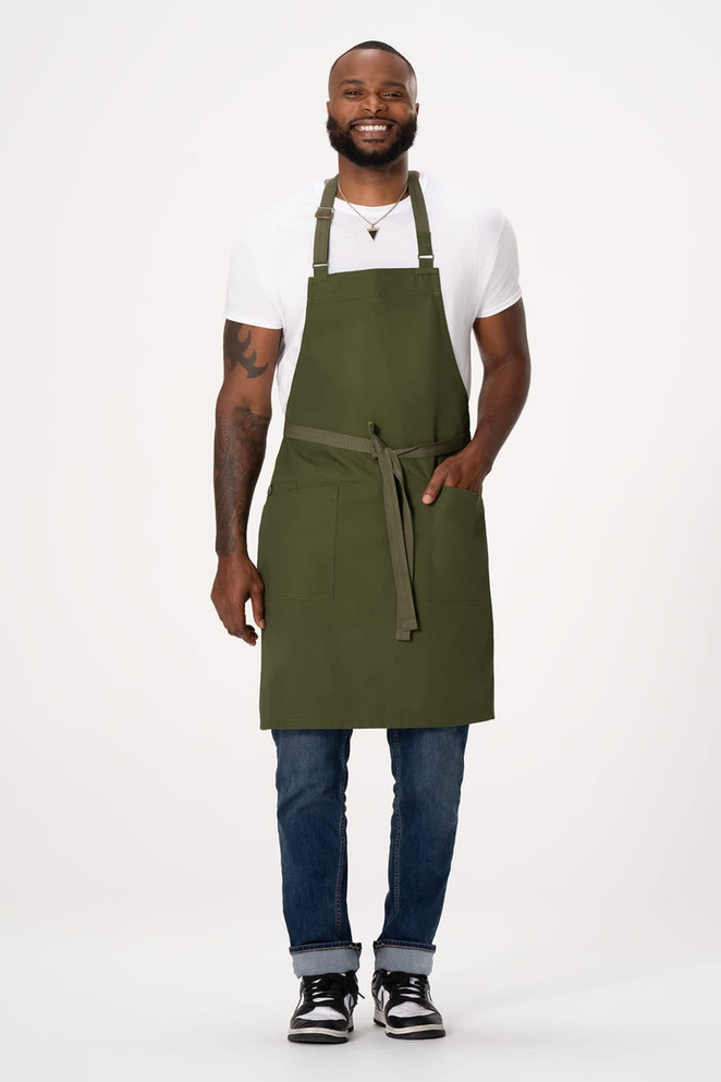 A man wearing a dark green Lockharte Bib Apron over a white t-shirt and jeans, with a front pocket and adjustable straps.