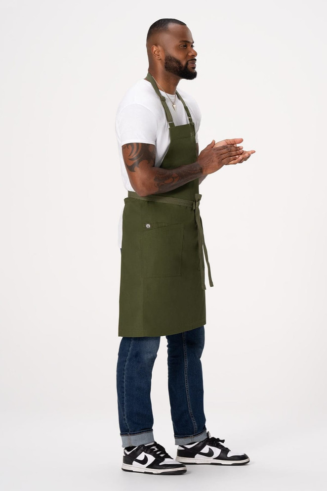 An olive green bib apron worn by a person, featuring a logo, with casual attire and sneakers.