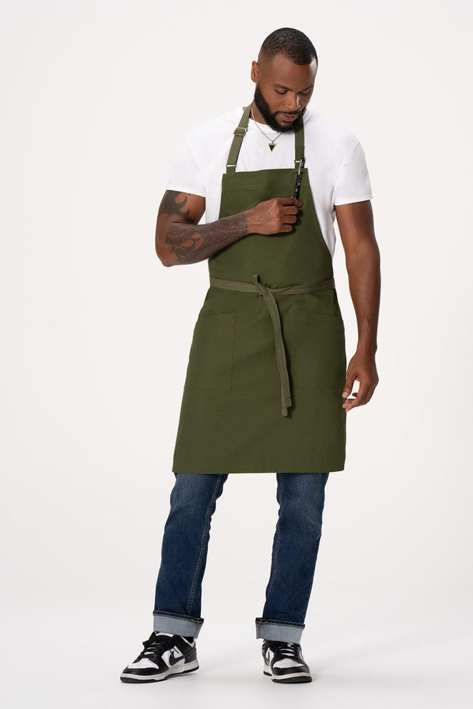 A man wearing an olive green Lockharte Bib Apron over a white T-shirt and jeans, displaying a front pocket.