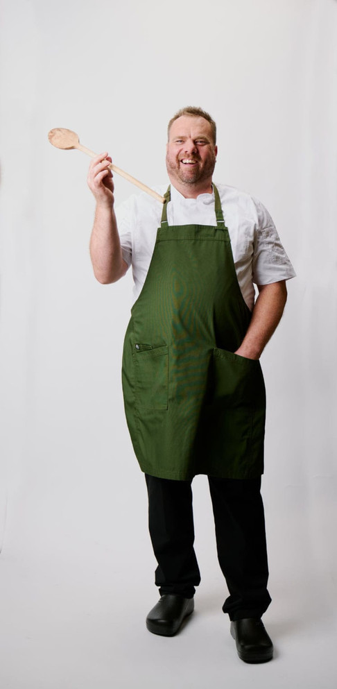 A man in a green Lockharte Bib Apron holds a wooden spoon, standing against a light background.