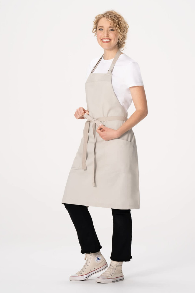 A light beige bib apron worn over a white t-shirt, tied at the waist, with a model standing and smiling.