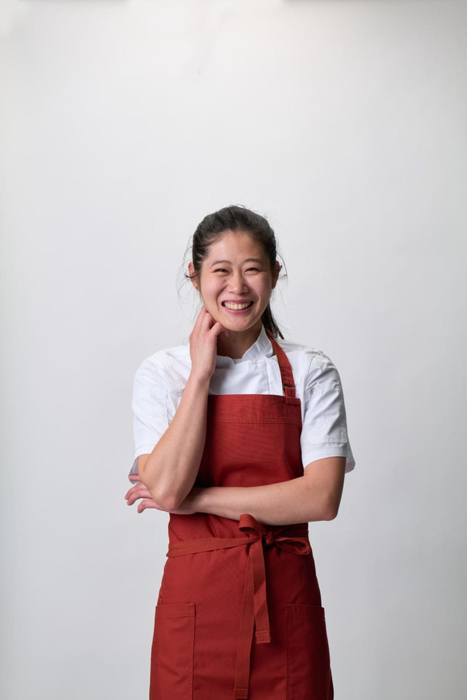 A smiling woman in a red bib apron stands against a plain background. The apron features a simple design.