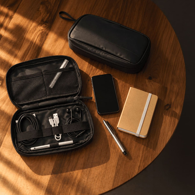 A black tech pouch containing cables, a pen, and a notebook, alongside a smartphone, on a wooden table.