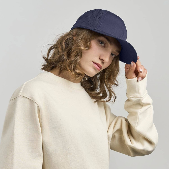 A woman wearing a navy cap and a cream-coloured sweatshirt, posing with one hand on her cap.